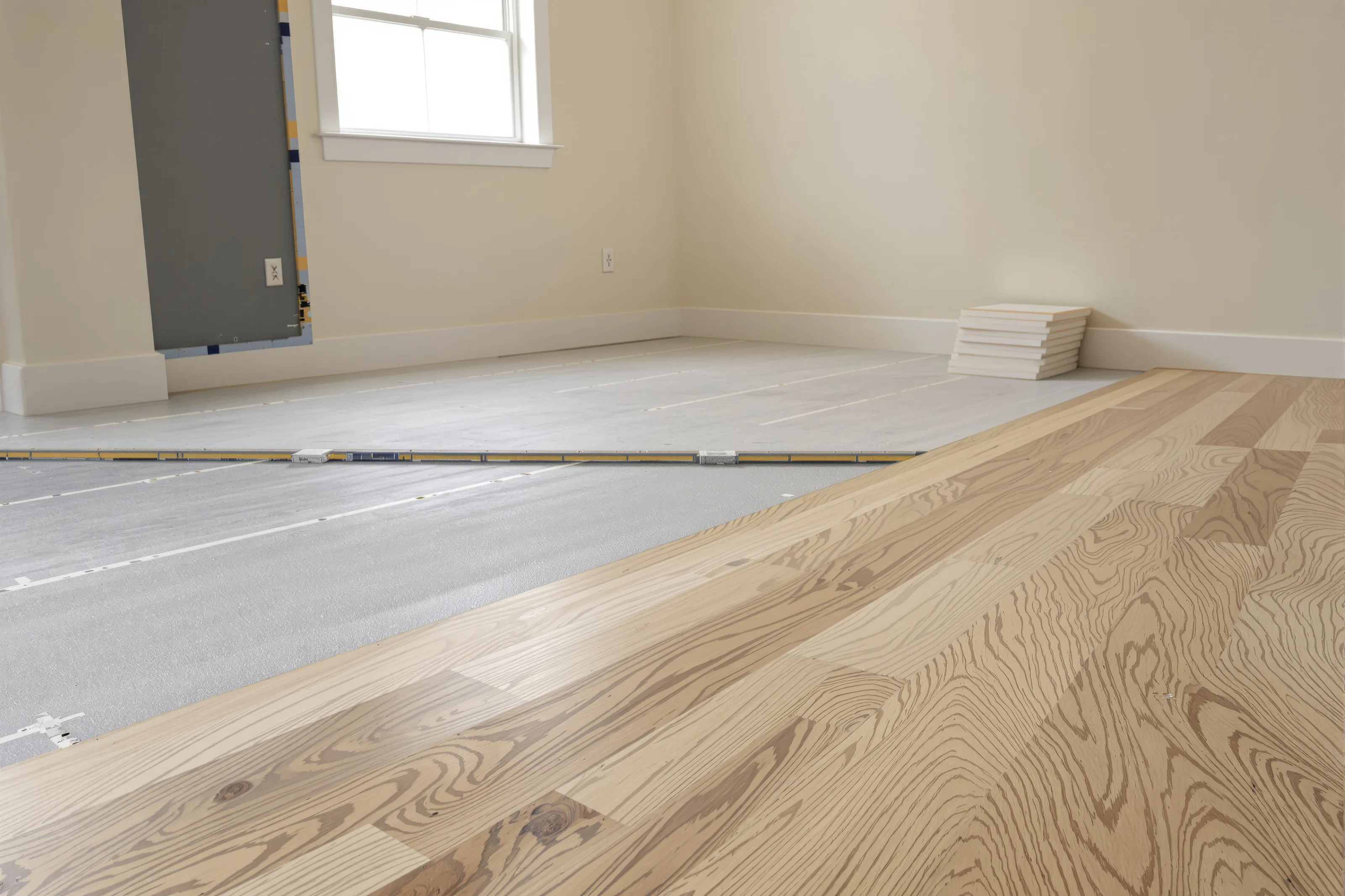 Empty room mid-renovation with wide-plank white oak half installed over grey underlayment and planks stacked by the wall